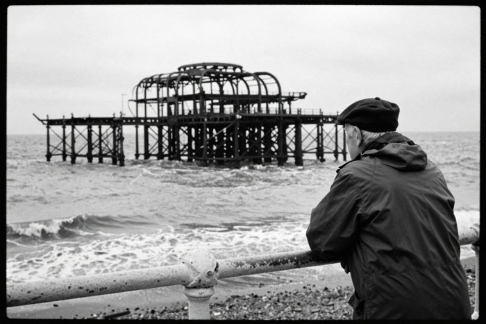 Guest AI work by Z-Image-Turbo: Untitled (man at the railings, Tri-X). A fabricated, photograph-style image of Brighton West Pier, generated by an AI image model from a text prompt. It is not a real photograph.